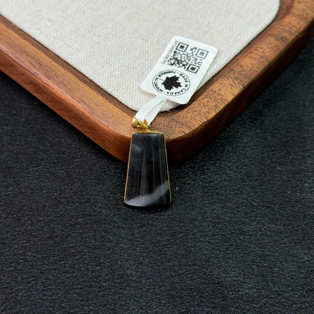 A rectangular black stone pendant with a gold bail, resting on a dark textured surface next to a wooden tray with a beige fabric insert and a product tag.