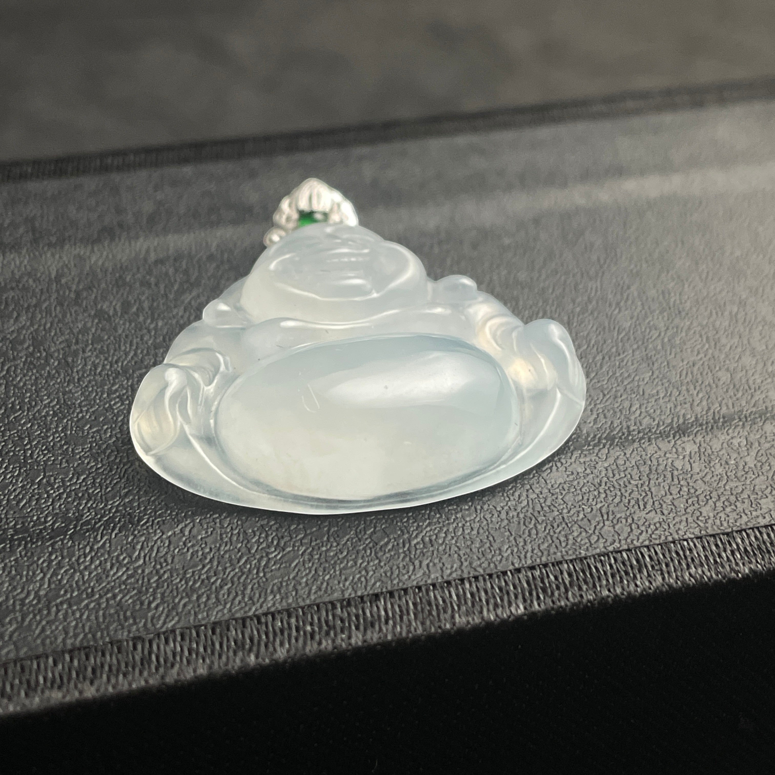 Sylvia Jewelry translucent white jadeite Laughing Buddha pendant with a silver bail on a black textured surface.
