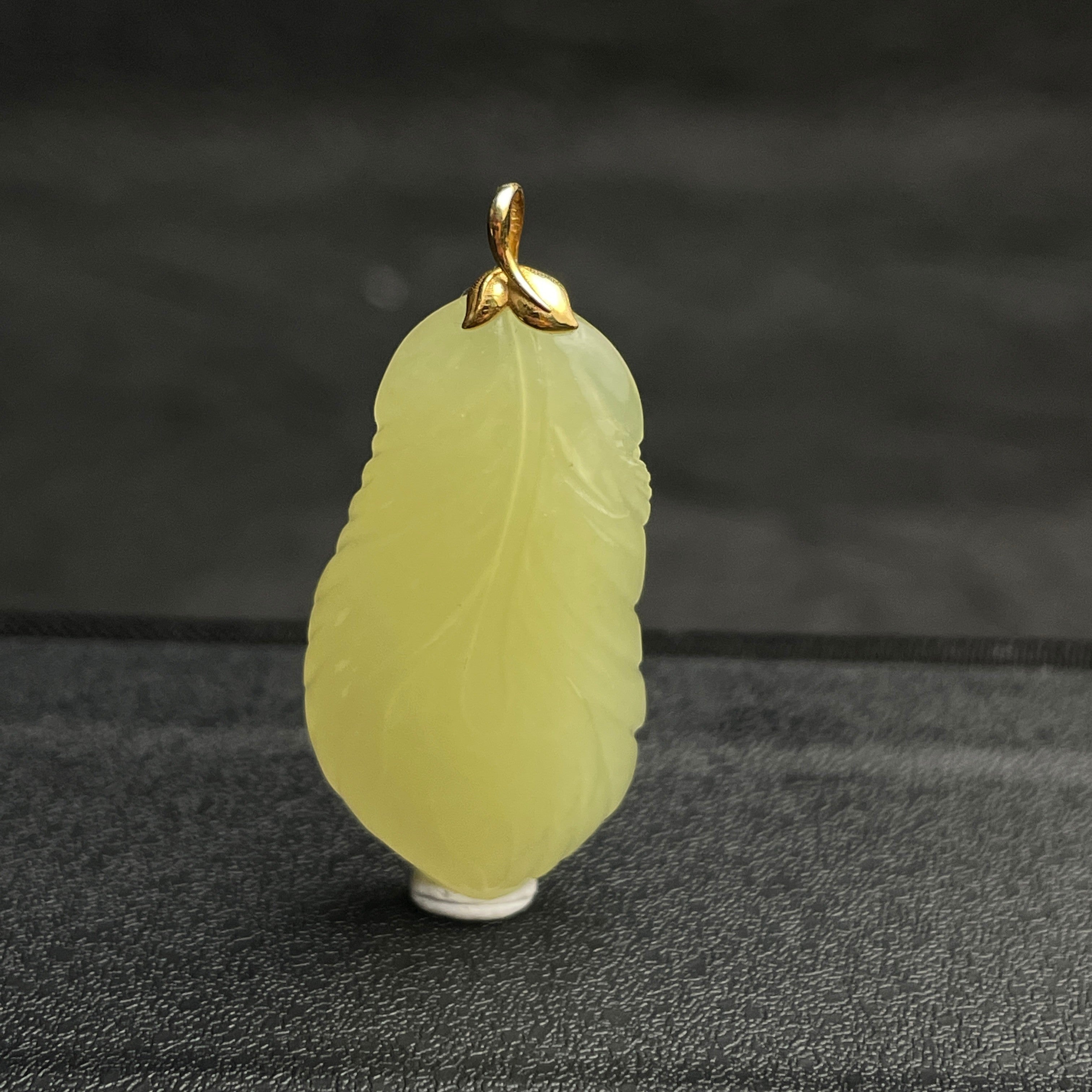 Sylvia Jewelry pale yellow jade feather pendant with a gold bail on a black textured background.