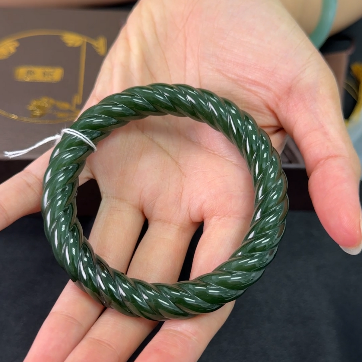 A dark green twisted bangle bracelet held in a person's hand, with a light blue jadeite bracelet visible on the wrist. The background is a mix of a beige top and a dark surface.