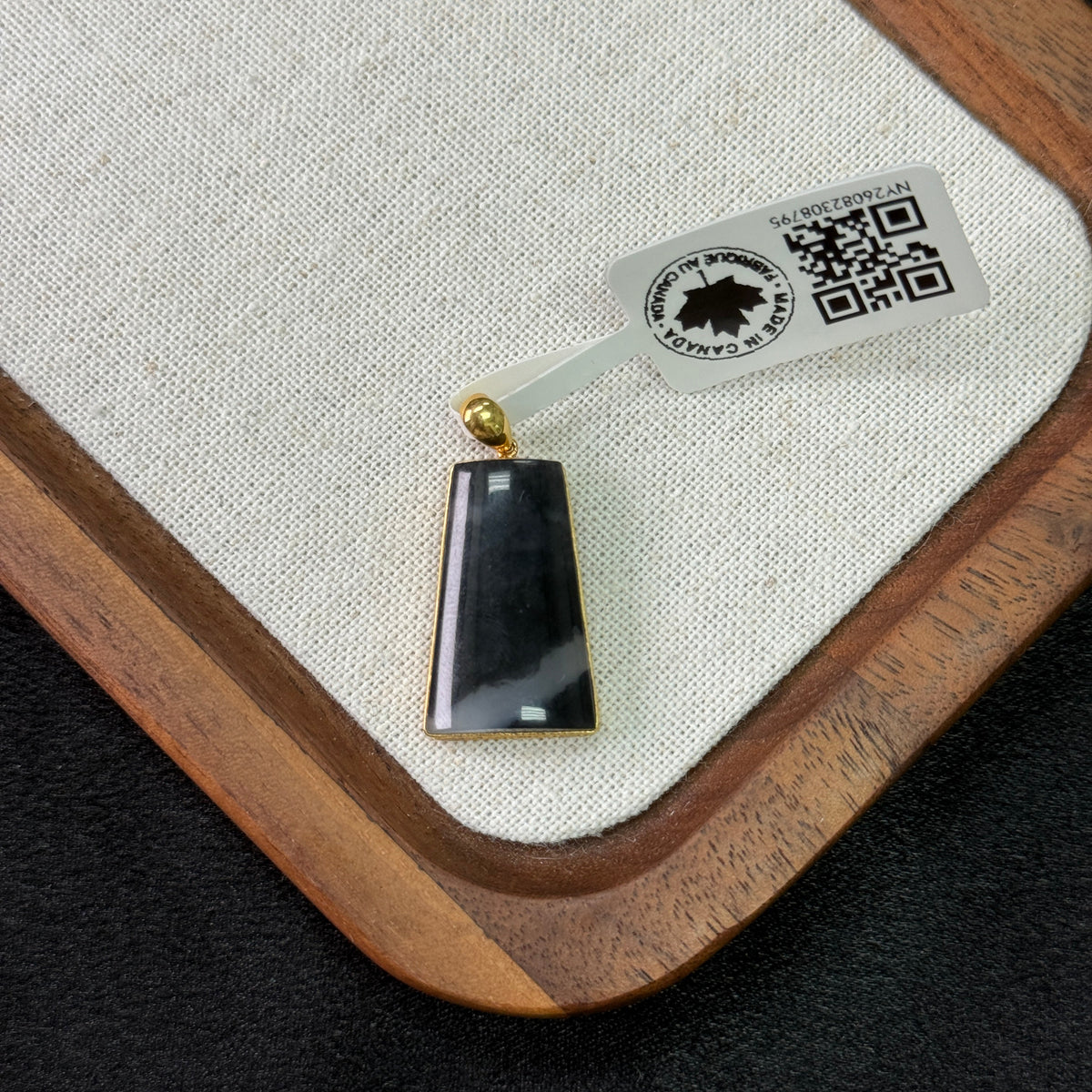 A rectangular black stone pendant with gold-toned metal borders and a gold bail, resting on a beige fabric-lined wooden tray with a product tag attached.