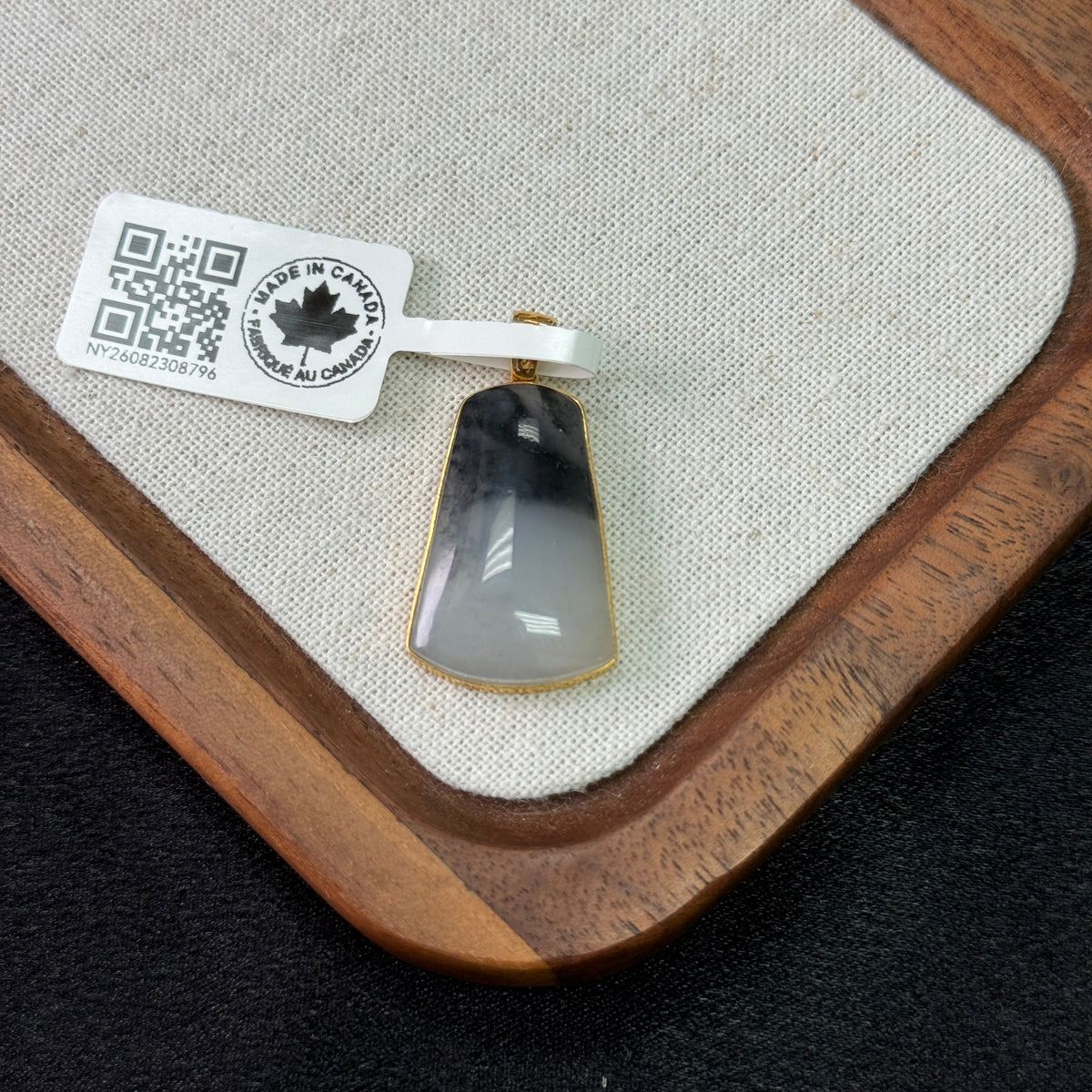 A teardrop-shaped pendant with a black and white marbled pattern, set in a gold-toned bezel, displayed on a beige fabric-lined wooden tray with a product tag attached.