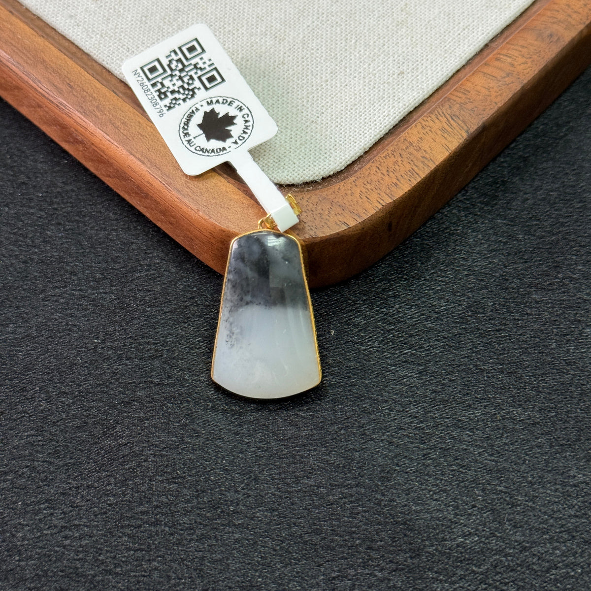 A teardrop-shaped pendant with a natural black and white marbled pattern, finished with a gold-toned bezel and hanging from a white tag, displayed on a dark textured surface next to a wooden tray.