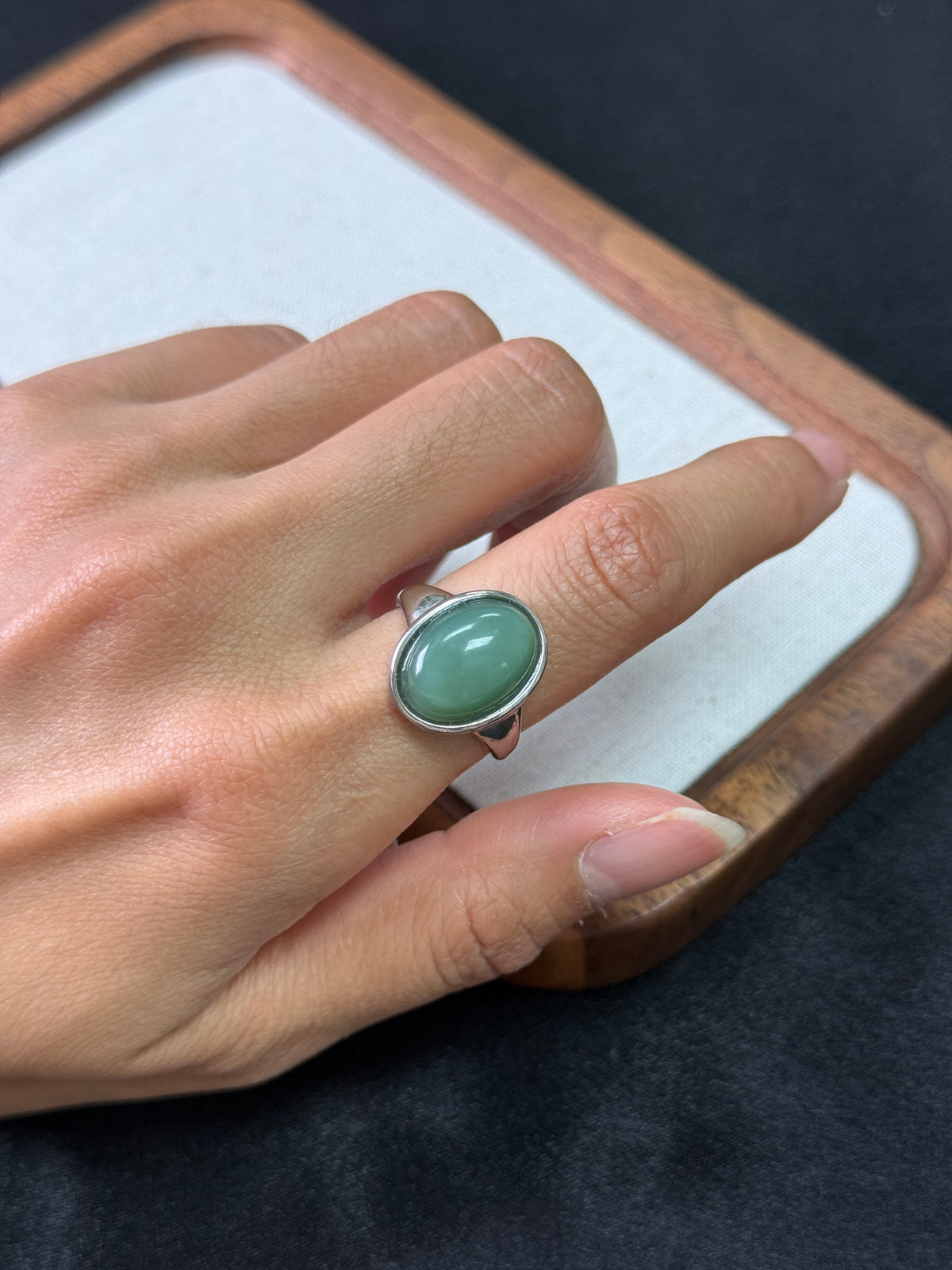 A close-up of a hand wearing a silver ring with an oval light green stone, resting on a white surface with a wooden frame.