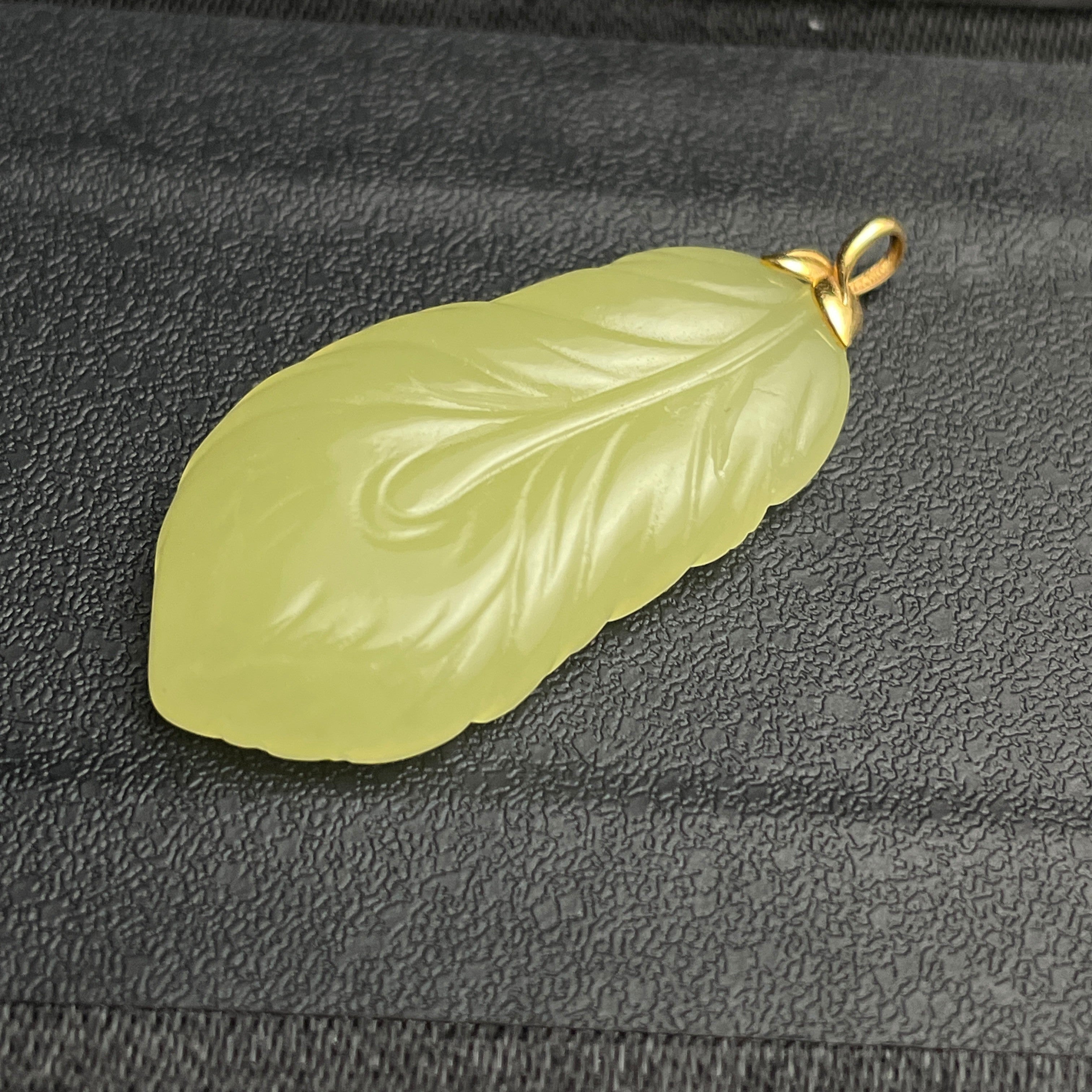 Sylvia Jewelry pale green jade feather pendant with a gold bail on a textured black background