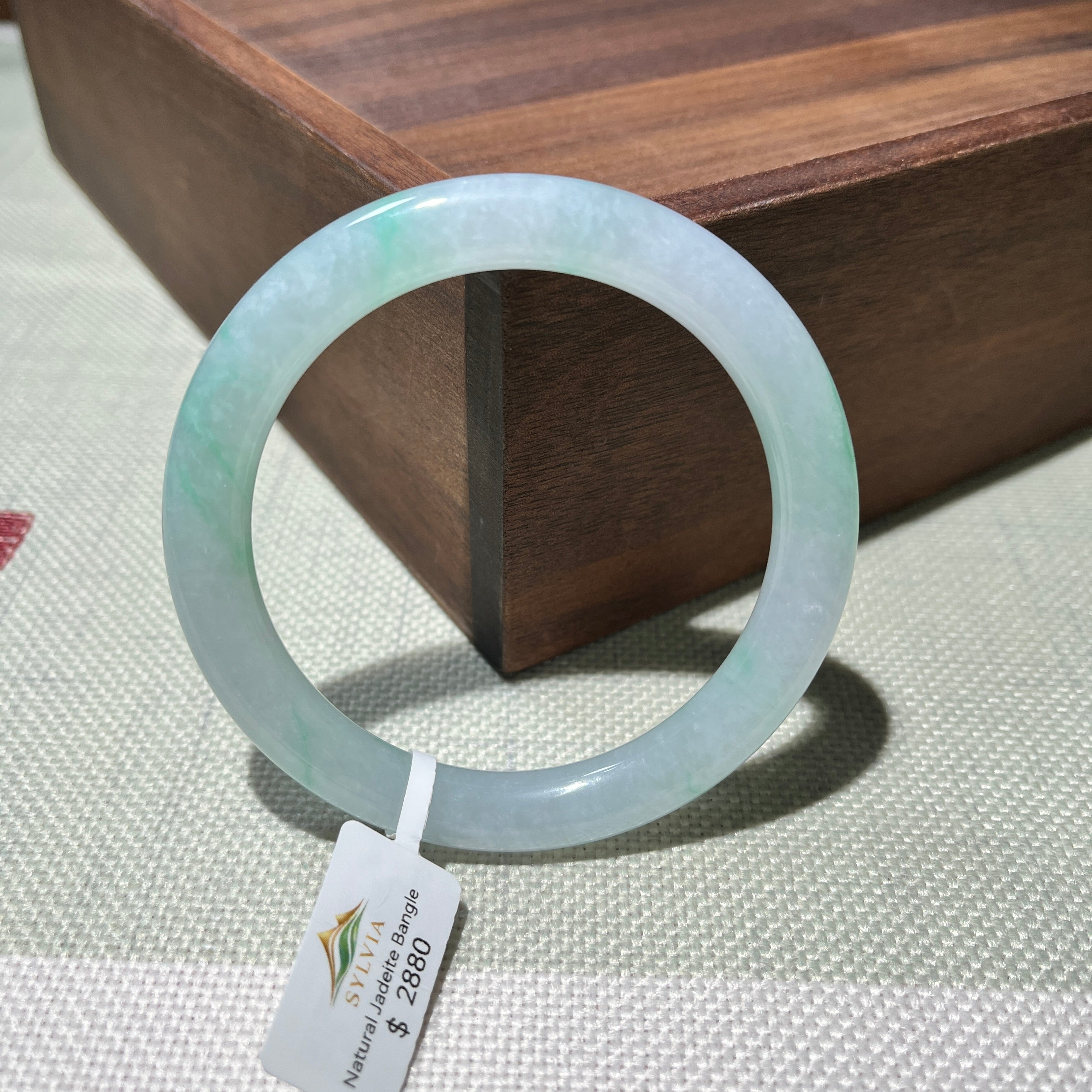 Natural jadeite bangle with an icy white body and soft spring green colour, photographed beside a wooden display box